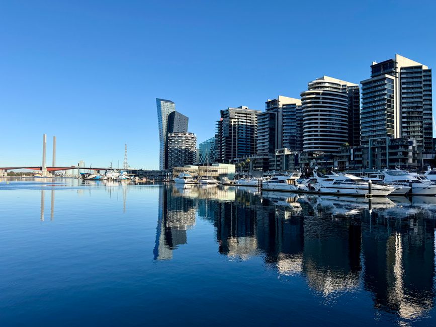 A panoramic view of a modern urban marina showing a collection of high-rise commercial and residential buildings with reflective glass exteriors, situated along a calm waterway under a clear blue sky. Several white yachts and motorboats are docked at the marina, their reflections visible on the smooth surface of the water. The foreground features a pier or quay with wooden posts, and the backdrop includes a cityscape with contemporary architecture. The lighting indicates a bright, sunny day, highlighting the textures of the buildings and the peaceful water. The scene exemplifies a developed waterfront area that may facilitate private boat moorings and city living, linking to the topic of alternative waste handling such as rubbish removal services in urban dockland locations.