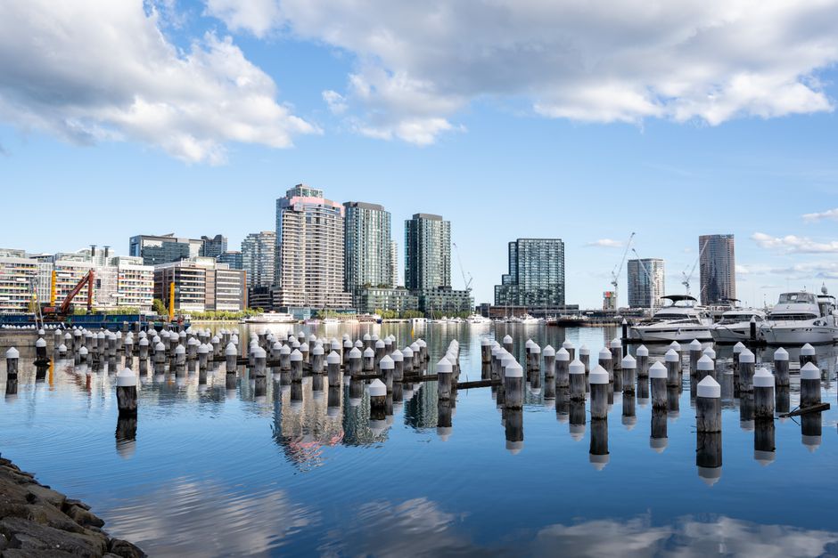 A waterfront scene in an urban area showing a calm body of water in the foreground, with a row of weathered wooden pylons lining the edge, each topped with a white cap. Beyond the water, there is a paved promenade with a line of evenly spaced small green trees, and behind the trees, modern high-rise buildings and a large sports stadium with a distinctive curved, steel-framed roof structure. The stadium's exterior features signage, including the number 7 and a red and black logo. The area is bathed in bright daylight, with blue sky and minimal cloud cover, creating a clear and vibrant atmosphere. The scene suggests a city centre riverside location, potentially used for events or leisure activities, with no visible rubbish or waste materials. The presence of the stadium and commercial buildings emphasizes the urban context relevant to alternative waste management and rubbish removal services in a busy metropolitan environment.