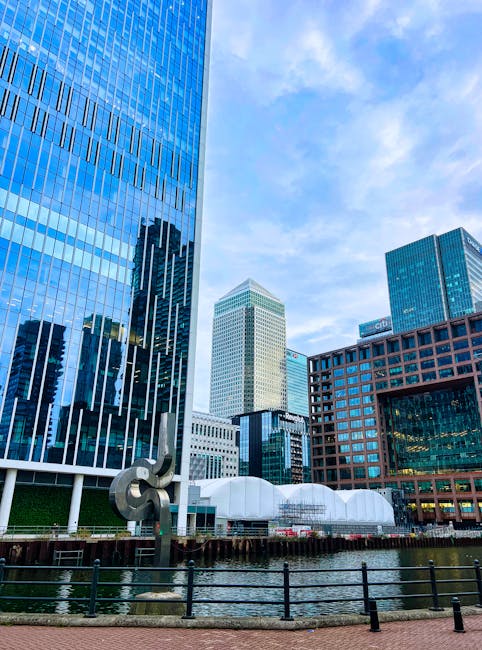 A daytime view of a modern cityscape featuring several tall office buildings with reflective glass facades, set along a canal or waterfront. The buildings include one with vertical blue-tinted windows on the left, another with a beige-colored grid pattern in the background, and a brick-red and glass structure on the right. In the foreground, a black metal railing runs along the edge of the water, which reflects parts of the buildings. Near the water, a large abstract metal sculpture with curved and intertwined forms is visible. Behind the sculpture, there are several white, semi-domed structures that appear to be temporary or portable with fabric or plastic coverings. The scene is illuminated by natural daylight, with a partly cloudy sky overhead, creating a bright but soft overall light that highlights the textures and colors of the buildings and waterfront area. The image broadly relates to urban environments where alternative waste management or rubbish removal services may operate, with the presence of the waterfront and modern infrastructure suggesting a setting suited for private and independent waste collection activities, such as those offered by Waste Removal Docklands.