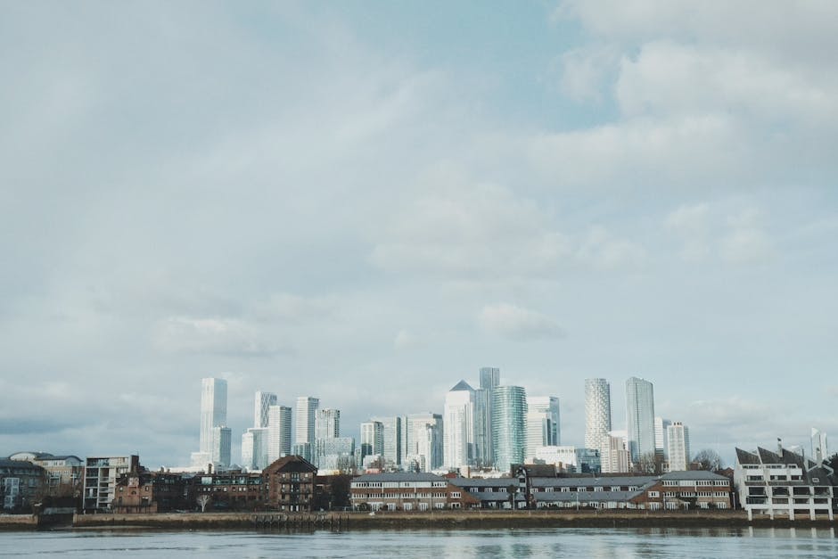 A distant city skyline featuring modern, glass-clad high-rise buildings of varying heights, with reflective surfaces showing the surrounding sky and clouds, situated behind a lower stretch of brick and concrete buildings along the waterfront. In the foreground, there is a body of water with a smooth, reflective surface, and the sky above is partly cloudy with soft, diffused lighting. The scene depicts an urban environment with a mix of architectural styles, from older structures near the water to contemporary skyscrapers, aligning with scenes where private waste removal services like Waste Removal Docklands might operate in the context of managing commercial and residential waste in city areas.