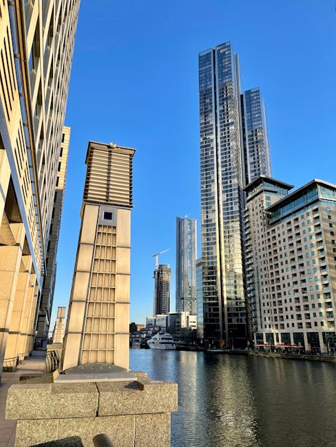 A daytime view of a modern cityscape featuring several tall office buildings with reflective glass facades, set along a canal or waterfront. The buildings include one with vertical blue-tinted windows on the left, another with a beige-colored grid pattern in the background, and a brick-red and glass structure on the right. In the foreground, a black metal railing runs along the edge of the water, which reflects parts of the buildings. Near the water, a large abstract metal sculpture with curved and intertwined forms is visible. Behind the sculpture, there are several white, semi-domed structures that appear to be temporary or portable with fabric or plastic coverings. The scene is illuminated by natural daylight, with a partly cloudy sky overhead, creating a bright but soft overall light that highlights the textures and colors of the buildings and waterfront area. The image broadly relates to urban environments where alternative waste management or rubbish removal services may operate, with the presence of the waterfront and modern infrastructure suggesting a setting suited for private and independent waste collection activities, such as those offered by Waste Removal Docklands.
