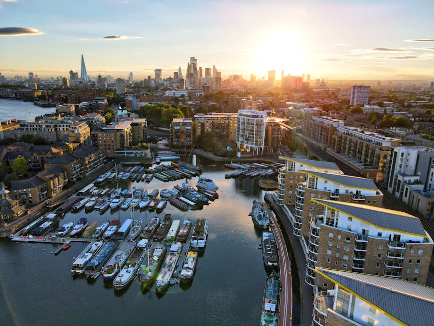 An aerial view of a modern marina in Docklands during sunset, with a variety of boats and yachts moored at the docks in the foreground, featuring sleek white hulls and polished surfaces. The water reflects the fading sunlight, creating a calm, shimmering surface. To the right, multi-storey residential buildings with contemporary glass balconies and light-colored facades are visible, lining the waterfront. Beyond, a mix of low-rise and high-rise buildings, including office towers and apartment complexes, extend towards the city skyline in the distance, highlighted by the warm glow of the setting sun and soft, scattered clouds overhead. The scene captures the urban landscape and waterway environment typical of a busy, regenerated dockside area. Waste Removal Docklands regularly handles waste clearance for such waterfront properties, supporting private on-site clearance or independent collection for rubbish that accumulates within these residential and commercial developments, emphasizing the importance of professional waste management in busy city environments.