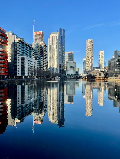 A panoramic view of a modern city skyline reflected in a calm river or canal, featuring high-rise buildings with glass and concrete facades, some with distinctive architectural designs such as a cylindrical tower and rectangular structures. The buildings are set against a clear blue sky with minimal cloud cover, and the water's surface creates a mirror-like reflection of the urban landscape. In the foreground, the water is smooth, with subtle ripples, and the edges of the riverbank are visible with some structures and possibly a bridge or walkway in the background. The natural lighting suggests daytime with bright, direct sunlight illuminating the scene. This urban environment, characterized by a concentration of tall buildings and a waterway, highlights the typical setting where local property owners or businesses might seek private waste management solutions, such as rubbish removal services from Waste Removal Docklands, rather than relying solely on municipal collection.