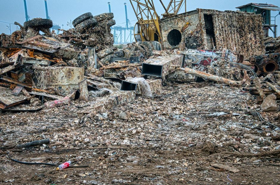 Image depicting a large pile of scrap metal and debris located outdoors, likely at a demolition or scrapyard site. The foreground is filled with various rusted metal pieces, including twisted sheets, pipes, and fragmented structural components, all coated in a layer of dirt and corrosion. A few black plastic containers and cylindrical objects are scattered among the debris. In the background, a yellow crane arm extends upward, indicating active dismantling or site clearance activities. The surrounding environment includes a mixture of muddy ground and scattered smaller metal fragments, with industrial structures and cranes visible against a cloudy sky. The scene reflects a typical setting for waste removal or private disposal of scrap materials, aligning with services offered by waste removal companies like Waste Removal Docklands, particularly for on-site clearance or alternative waste handling outside conventional recycling centers.