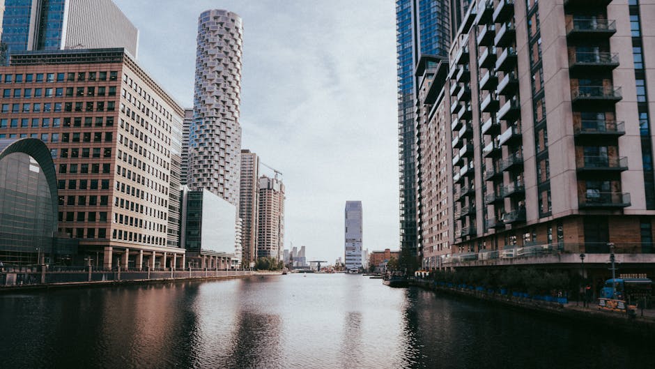 A wide view of a cityscape with tall modern skyscrapers lining a river or canal, the buildings feature a variety of materials including glass, steel, and brick, with some having balconies and reflective surfaces. The water in the foreground appears calm, with slight ripples, and reflects the surrounding structures. The sky is partly cloudy, casting diffuse daylight over the scene. On the left side, there is a prominent curved building with a dark facade, and on the right, a taller residential or office building with balconies and large windows. In the background, additional high-rise buildings and cranes indicate ongoing construction. The environment is urban, with the river acting as a central feature amidst dense development. This scene is consistent with city areas where urban renewal or private waste management services, such as those by Waste Removal Docklands, might be relevant for handling rubbish or waste collection related to high-density properties, visible through absence of waste containers but contextual to the service focus.