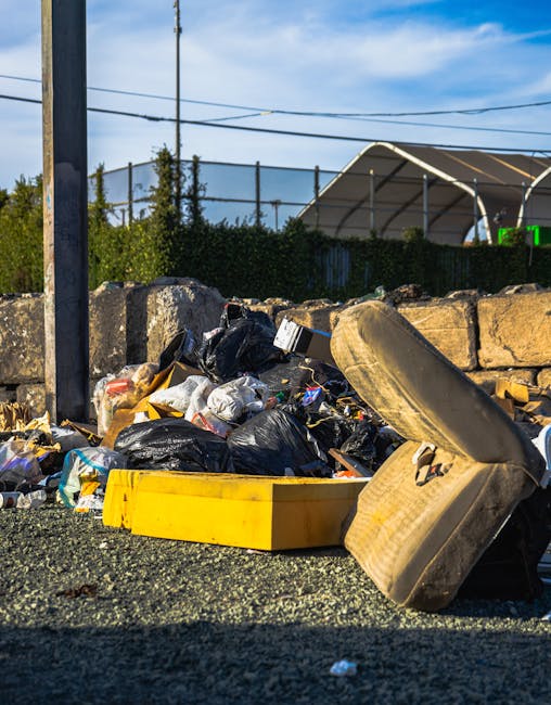 Image depicting a large pile of scrap metal and debris located outdoors, likely at a demolition or scrapyard site. The foreground is filled with various rusted metal pieces, including twisted sheets, pipes, and fragmented structural components, all coated in a layer of dirt and corrosion. A few black plastic containers and cylindrical objects are scattered among the debris. In the background, a yellow crane arm extends upward, indicating active dismantling or site clearance activities. The surrounding environment includes a mixture of muddy ground and scattered smaller metal fragments, with industrial structures and cranes visible against a cloudy sky. The scene reflects a typical setting for waste removal or private disposal of scrap materials, aligning with services offered by waste removal companies like Waste Removal Docklands, particularly for on-site clearance or alternative waste handling outside conventional recycling centers.
